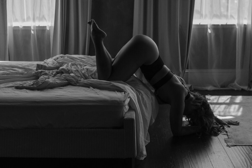 Black and white boudoir portrait of woman arching over bed with dramatic shadows